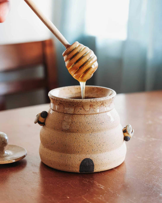Honey dipper drizzling honey into a ceramic jar on a wooden table.